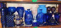 Shelf display of assorted cobalt blue glassware including bottles, candlesticks, and vases along with ceramic and stained glass vases