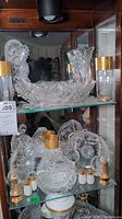 Cabinet shelf with various cut crystal candy dishes and cake slicer. Four gold-rimmed glasses and three sets of lusterware salt and pepper shakers placed in front on lower shelf.