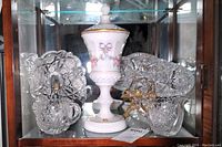 Photo inside a glass cabinet showing Westmoreland milk glass compote at center, surrounded by clear cut crystal bowls and pitchers with intricate patterns.