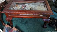 Top downward view of rectangular coffee table showing glass top inset and carved wood trim around edges. Inside display area visible with lace and small items beneath glass.