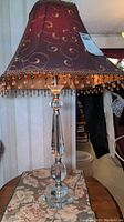 Full view of lamp showing burgundy silk shade embroidered with gold swirls and beaded fringe, along with clear faceted glass base on floral fabric surface.