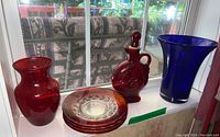 Photo showing ruby glass vase, ruby glass decanter with stopper, three ruby glass plates and cobalt blue glass vase on a window ledge.