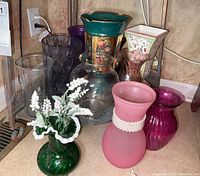 Nine assorted glass and ceramic vases in various colors and styles arranged on floor.