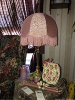 Photo showing Victorian style table lamp with pink floral fabric lampshade and brass base standing on floor beside floral upholstered armchair. Pink glass swan candle holders, pink candles and Royal Vinton floral plate are on table at lamp base.