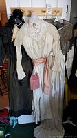Full view of vintage and antique Victorian clothing, showing cream dress with lace collar and pink sash, dark dress with feathered collar, plus additional dresses and accessories.