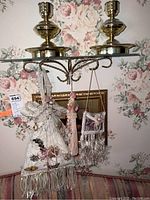Two brass candle holders on a glass shelf with floral wallpaper background, several decorative pins hanging.