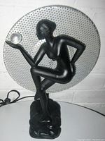 Front view of black figural nude lady lamp with clear glass orb and silver perforated backdrop on white surface.