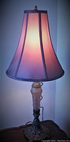 Full view of the lit lamp showing the lilac pink glass base and the matching purple multi-panel fabric shade glowing softly.