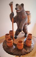 Front and slightly side angled view of the carved wooden bear standing on a carved base with six wooden shot cups placed around it. Bear carrying a barrel and holding a horn.
