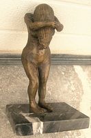 Front angle view of the bronze nude statue mounted on black marble base showing figure covering face with hair and arms.