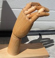 Wood carved articulated hand with bent fingers shown from side angle casting shadow on white wall, mounted on rectangular wood base.