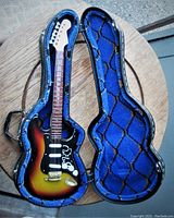 Open case with miniature electric guitar inside, showing front with sunburst finish, white pickguard with 'IRV' lettering, and knobs