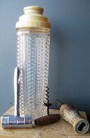 Clear pressed glass Art Deco cocktail shaker with cream plastic cap, next to chrome bottle opener, wooden handled corkscrew, and novelty duck head bottle opener