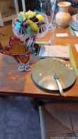 Wide shot showing amber Murano vase, faux fruit in footed glass bowl, marble cheese board with wire slicer, and round green marble charcuterie board with silver spreader