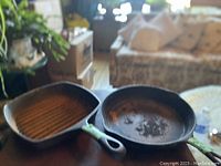 Photo showing two cast iron pans, one round frying pan and one square grill pan, both with visible wear and patina.