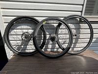Three bicycle wheels standing vertically against siding and placed on a wooden surface, showing front side of wheels.