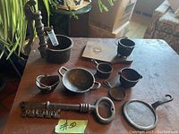 Photo shows nine small cast iron items on a wooden table, including miniature pans, pots, a ladle, small cups, and a decorative key-shaped cast iron piece with the word 'CALGARY' on it.