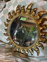 Mirror with gilded wooden flame-styled frame lying on quilt, showing overall condition and style.
