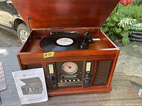 Full view of wood cabinet music center with hinged lid open showing vinyl record on turntable and instruction manual on side.