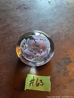Glass paperweight with visible white floral design inside, placed on a wooden surface with label '#63' nearby.