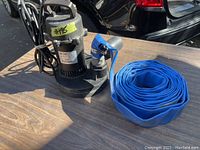 Full view of the BurCam submersible sump pump on a wood surface with coiled blue discharge hose attached and power cord bundled on the side.