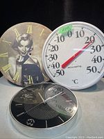 Front view of three round items: Marlene Dietrich art clock with Roman numerals, white AcuRite outdoor thermometer, and black quartz wall clock.