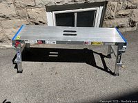 Front view of aluminum folding work bench with visible side leg supports and surface with aluminum slats and blue end caps.