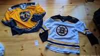 Both Boston Bruins hockey jerseys side by side on wooden floor, yellow jersey on left, white jersey on right