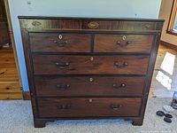 Full front view of five-drawer dresser showing wood grain, inlaid shell motifs on top rail, brass pulls and escutcheons