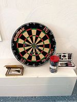 Full setup including Winmau Blade 4 dartboard, multiple darts, dart accessories, and storage container on a white surface.