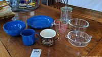 Full view showing four blue enamelware bowls, four blue enamelware plates, one blue enamelware mug, one brown Howler Bros mug, Pyrex measuring cup, large glass bowl, glass bowl with handles, and a glass canning jar