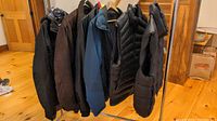 Rack with six men's coats and jackets hung in a row, dark color palette.