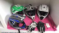 Four motocross helmets and two pairs of gloves arranged on a pink cloth, front view showing helmet designs and gloves.