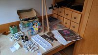 A view of many acrylic paint tubes, palettes, brushes, easels, and sketch pads arranged on a table against a wooden wainscot wall.