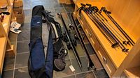 Photo showing two pairs of skis with bindings, ski poles, and two ski bags on tiled floor.