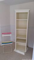 Front view of white swivel bookcase next to rainbow folding chair.