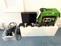 Front view of Xbox One console, four wireless controllers, original box, and a box of various cables and accessories.