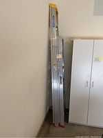 Two aluminum extension ladders leaning against a wall with white cabinet beside them. One ladder is approximately 8 ft, the other 6 ft. Both have yellow caps at top and red rubber feet at base.