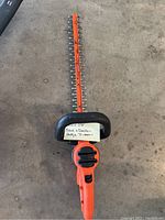 Top-down photo of the entire hedge trimmer showing orange body, blade with teeth, and black protective guard with a label.