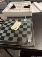 Glass chess board with frosted and clear squares, clear bishop and frosted pawn pieces on board. Partial styrofoam packaging visible in background.