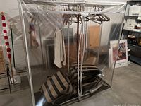 Fully enclosed metal clothing rack with clear plastic vinyl cover and metal frame with hangers inside.