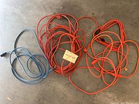 Four coiled extension cords on concrete floor: two orange cords and two blue cords, varying lengths and outlet types.