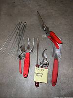 Photo showing small gardening hand tools arranged on concrete floor including pruning shears, rake, shovel, garden fork, and metal stakes.