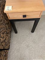 Front view of one wooden nightstand showing light wood drawer with black knob and dark legs beside bed