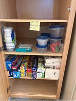 Photo showing plastic food storage bowls with blue lids, disposable paper cups in white with patterns, plastic bags, and napkins arranged in a cabinet.