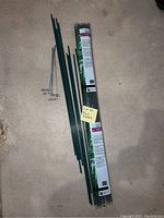 Green plant stakes laid out with some metal wire supports visible, next to a rolled package of plant stakes labeled 'Plant Stakes'.