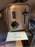 Front view of retro toaster showing brushed metal finish, three function buttons (bagel, defrost, reheat), and rotary dial.