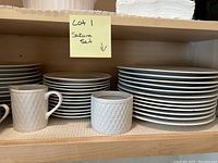 Shelf holding stacked large and small plates, mugs, and a small container with basketweave texture.