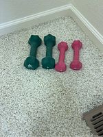 Four small hand weights displayed on carpeted floor, two green and two pink, side by side.
