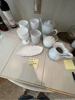 White ceramic goblets, bowls, leaf-shaped dish, creamer and teapots displayed on tile countertop. Items need cleaning, shown from top-down.